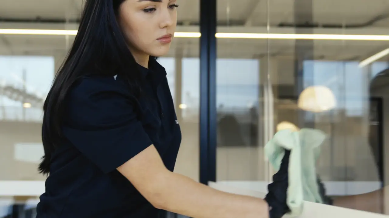 A Castle Cleans cleaner wiping a glass partition in an office, with workstations visible behind